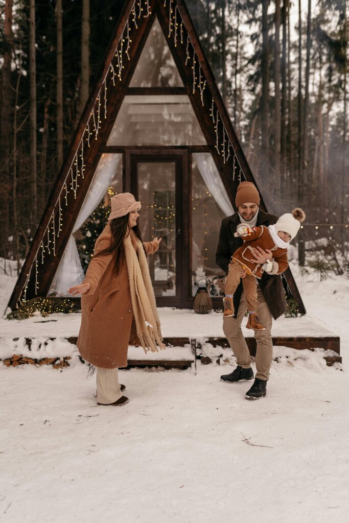 A joyful family playing in the snow outside a decorated A-frame cabin in the forest.