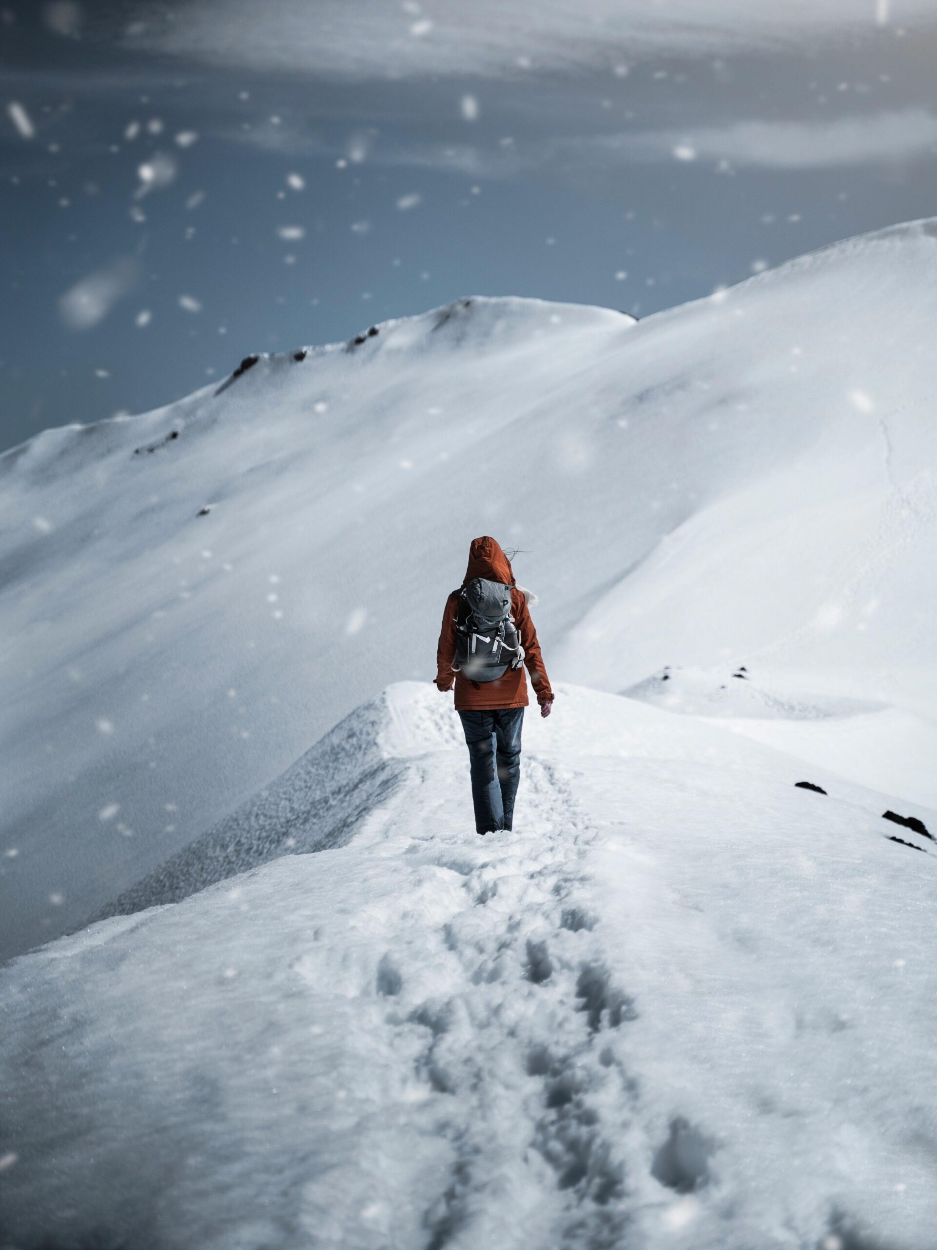 A lone hiker in a red jacket walks to explore nature on a snow-covered mountain path during a snowfall.
