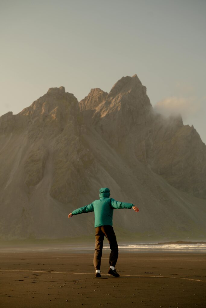 Person in a teal jacket poses on a black sand beach in Iceland, with mountains for exploring nature.
